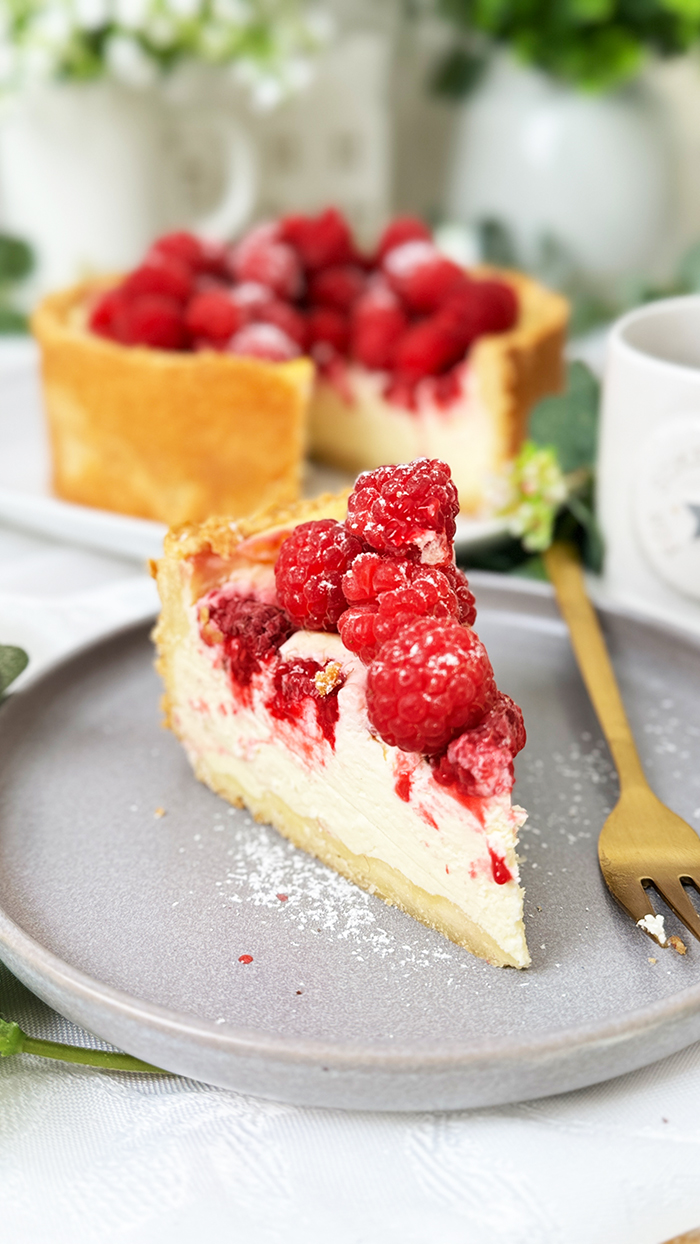 ein Stück Himbeer-Schmand-Kuchen auf einem Teller, garniert mit frischen Himbeeren und leicht mit Puderzucker bestäubt.