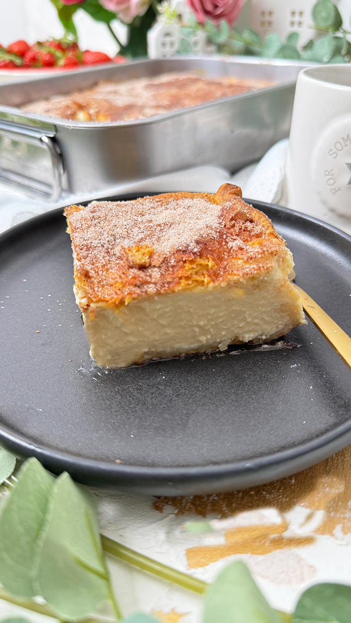 Churros Cheesecake-Schnitte auf einem schwarzen Teller mit einer goldenen Gabel. Die Schnitte hat eine goldbraune, knusprige Zimt-Zucker-Kruste und eine cremige Käsekuchenfüllung. Im Hintergrund steht eine Backform mit dem restlichen Kuchen sowie eine Schüssel mit frischen Erdbeeren und dekorativen Blumen