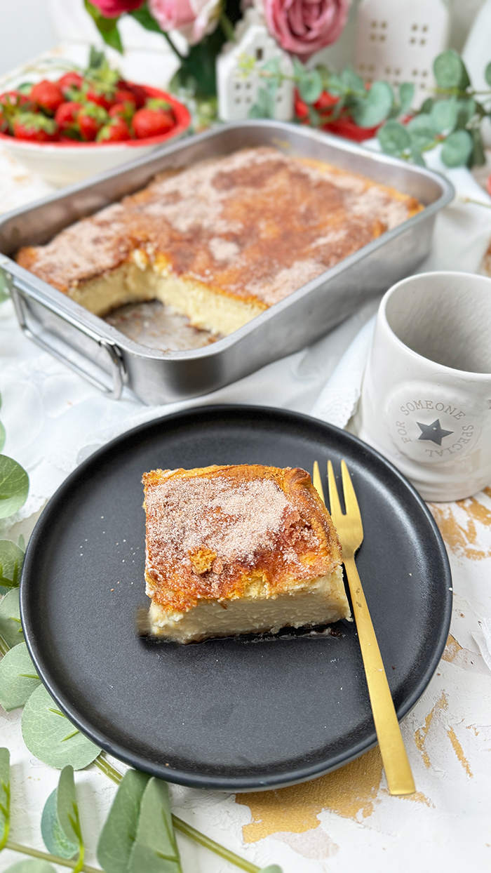 Churros Cheesecake-Schnitte auf einem schwarzen Teller mit einer goldenen Gabel. Die Schnitte hat eine goldbraune, knusprige Zimt-Zucker-Kruste und eine cremige Käsekuchenfüllung. Im Hintergrund steht eine Backform mit dem restlichen Kuchen sowie eine Schüssel mit frischen Erdbeeren und dekorativen Blumen