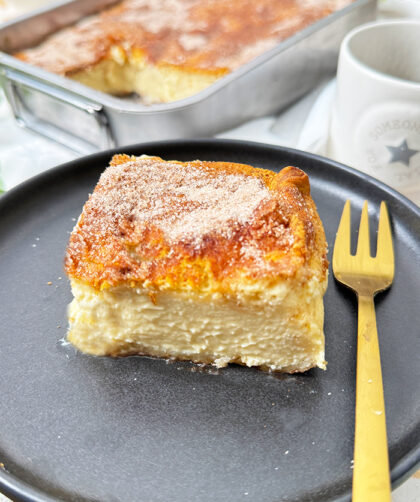 Churros Cheesecake-Schnitte auf einem schwarzen Teller mit einer goldenen Gabel. Die Schnitte hat eine goldbraune, knusprige Zimt-Zucker-Kruste und eine cremige Käsekuchenfüllung. Im Hintergrund steht eine Backform mit dem restlichen Kuchen sowie eine Schüssel mit frischen Erdbeeren und dekorativen Blumen