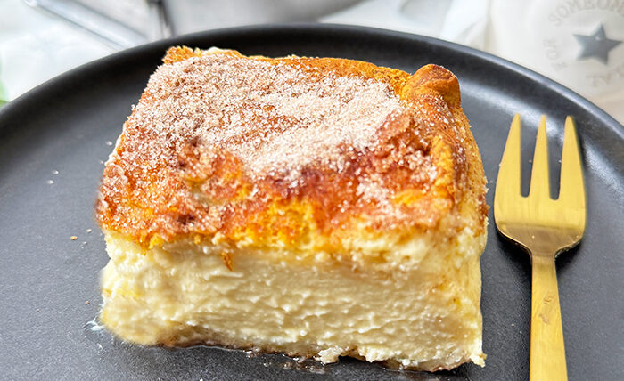 Churros Cheesecake-Schnitte auf einem schwarzen Teller mit einer goldenen Gabel. Die Schnitte hat eine goldbraune, knusprige Zimt-Zucker-Kruste und eine cremige Käsekuchenfüllung. Im Hintergrund steht eine Backform mit dem restlichen Kuchen sowie eine Schüssel mit frischen Erdbeeren und dekorativen Blumen