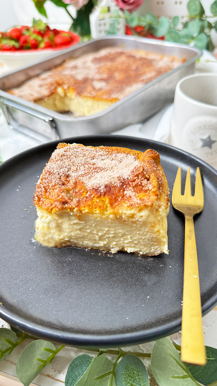 Churros Cheesecake-Schnitte auf einem schwarzen Teller mit einer goldenen Gabel. Die Schnitte hat eine goldbraune, knusprige Zimt-Zucker-Kruste und eine cremige Käsekuchenfüllung. Im Hintergrund steht eine Backform mit dem restlichen Kuchen sowie eine Schüssel mit frischen Erdbeeren und dekorativen Blumen