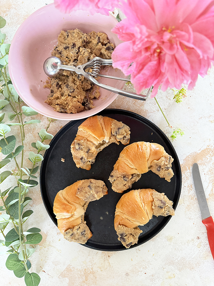 Das Bild zeigt vier Crookies auf einem schwarzen Teller, umgeben von rosa Blumen und Eukalyptuszweigen. Ein Crookie ist eine Kombination aus einem Croissant und einem Cookie. Die Crookies sind halb Croissant mit ihrem goldbraunen, glänzenden Teig und halb Cookie mit ihrem dickeren, bräunlichen Keksanteil, der Schokoladenstücke enthält.