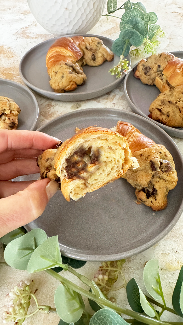 Das Bild zeigt vier Crookies auf einem schwarzen Teller, umgeben von rosa Blumen und Eukalyptuszweigen. Ein Crookie ist eine Kombination aus einem Croissant und einem Cookie. Die Crookies sind halb Croissant mit ihrem goldbraunen, glänzenden Teig und halb Cookie mit ihrem dickeren, bräunlichen Keksanteil, der Schokoladenstücke enthält.