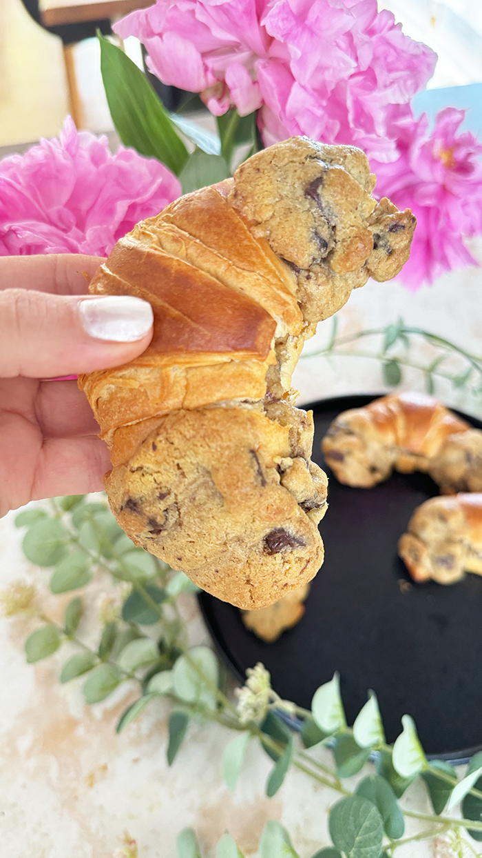 Das Bild zeigt vier Crookies auf einem schwarzen Teller, umgeben von rosa Blumen und Eukalyptuszweigen. Ein Crookie ist eine Kombination aus einem Croissant und einem Cookie. Die Crookies sind halb Croissant mit ihrem goldbraunen, glänzenden Teig und halb Cookie mit ihrem dickeren, bräunlichen Keksanteil, der Schokoladenstücke enthält.