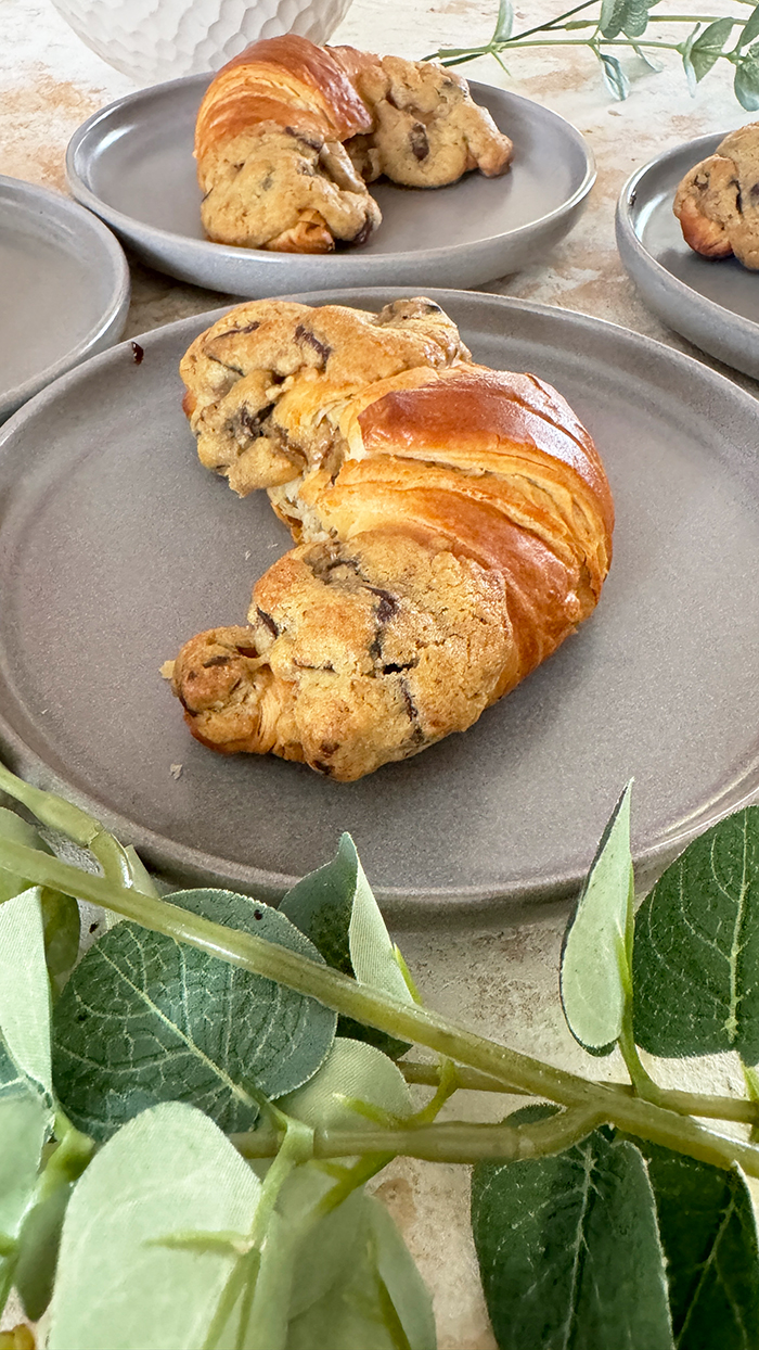 Das Bild zeigt vier Crookies auf einem schwarzen Teller, umgeben von rosa Blumen und Eukalyptuszweigen. Ein Crookie ist eine Kombination aus einem Croissant und einem Cookie. Die Crookies sind halb Croissant mit ihrem goldbraunen, glänzenden Teig und halb Cookie mit ihrem dickeren, bräunlichen Keksanteil, der Schokoladenstücke enthält.