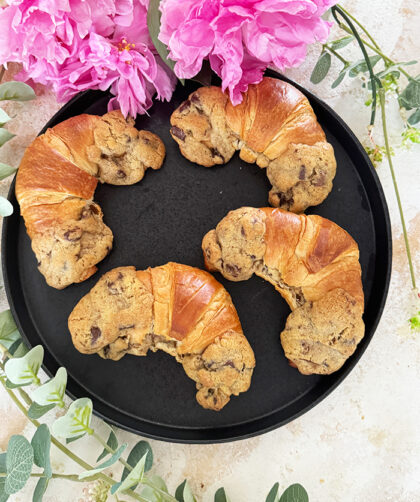 Das Bild zeigt vier Crookies auf einem schwarzen Teller, umgeben von rosa Blumen und Eukalyptuszweigen. Ein Crookie ist eine Kombination aus einem Croissant und einem Cookie. Die Crookies sind halb Croissant mit ihrem goldbraunen, glänzenden Teig und halb Cookie mit ihrem dickeren, bräunlichen Keksanteil, der Schokoladenstücke enthält.