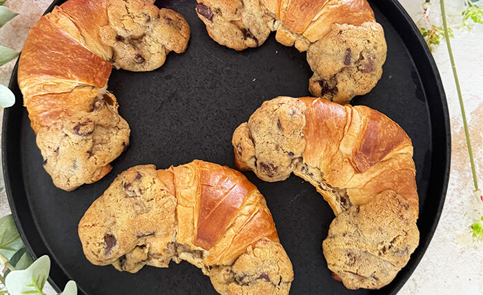 Das Bild zeigt vier Crookies auf einem schwarzen Teller, umgeben von rosa Blumen und Eukalyptuszweigen. Ein Crookie ist eine Kombination aus einem Croissant und einem Cookie. Die Crookies sind halb Croissant mit ihrem goldbraunen, glänzenden Teig und halb Cookie mit ihrem dickeren, bräunlichen Keksanteil, der Schokoladenstücke enthält.