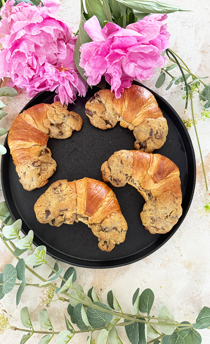 Das Bild zeigt vier Crookies auf einem schwarzen Teller, umgeben von rosa Blumen und Eukalyptuszweigen. Ein Crookie ist eine Kombination aus einem Croissant und einem Cookie. Die Crookies sind halb Croissant mit ihrem goldbraunen, glänzenden Teig und halb Cookie mit ihrem dickeren, bräunlichen Keksanteil, der Schokoladenstücke enthält.