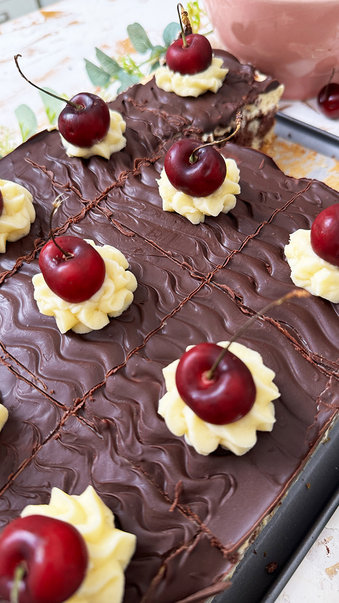 Ein köstliches Blech Donauwelle-Kuchen, dekoriert mit Sahnetupfern und frischen Kirschen. Ein einzelnes Stück des Kuchens liegt auf einem schwarzen Teller mit einer goldenen Gabel. Im Hintergrund befinden sich eine Schüssel voller Kirschen, rosafarbene Blumen und kleine weiße Dekohäuschen. Der Kuchen zeigt die typischen Schichten von hellem und dunklem Teig, Buttercreme und einer Schokoladenglasur