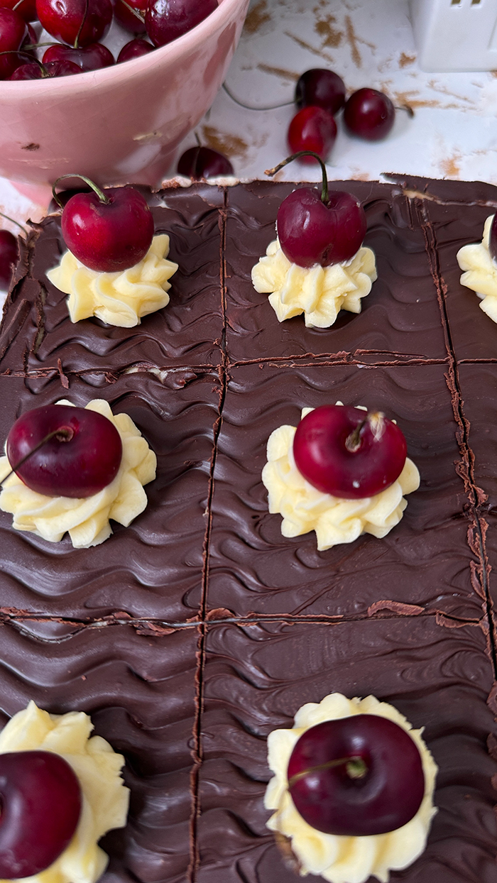 Ein köstliches Blech Donauwelle-Kuchen, dekoriert mit Sahnetupfern und frischen Kirschen. Ein einzelnes Stück des Kuchens liegt auf einem schwarzen Teller mit einer goldenen Gabel. Im Hintergrund befinden sich eine Schüssel voller Kirschen, rosafarbene Blumen und kleine weiße Dekohäuschen. Der Kuchen zeigt die typischen Schichten von hellem und dunklem Teig, Buttercreme und einer Schokoladenglasur