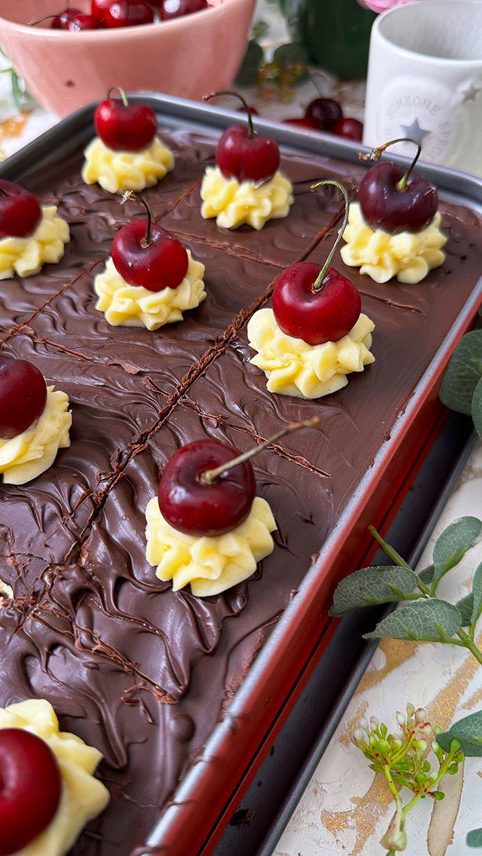 Ein köstliches Blech Donauwelle-Kuchen, dekoriert mit Sahnetupfern und frischen Kirschen. Ein einzelnes Stück des Kuchens liegt auf einem schwarzen Teller mit einer goldenen Gabel. Im Hintergrund befinden sich eine Schüssel voller Kirschen, rosafarbene Blumen und kleine weiße Dekohäuschen. Der Kuchen zeigt die typischen Schichten von hellem und dunklem Teig, Buttercreme und einer Schokoladenglasur