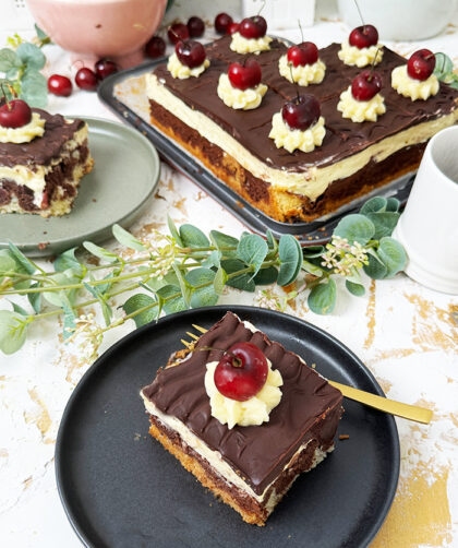 Ein köstliches Blech Donauwelle-Kuchen, dekoriert mit Sahnetupfern und frischen Kirschen. Ein einzelnes Stück des Kuchens liegt auf einem schwarzen Teller mit einer goldenen Gabel. Im Hintergrund befinden sich eine Schüssel voller Kirschen, rosafarbene Blumen und kleine weiße Dekohäuschen. Der Kuchen zeigt die typischen Schichten von hellem und dunklem Teig, Buttercreme und einer Schokoladenglasur