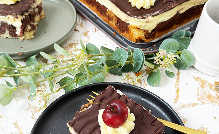 Ein köstliches Blech Donauwelle-Kuchen, dekoriert mit Sahnetupfern und frischen Kirschen. Ein einzelnes Stück des Kuchens liegt auf einem schwarzen Teller mit einer goldenen Gabel. Im Hintergrund befinden sich eine Schüssel voller Kirschen, rosafarbene Blumen und kleine weiße Dekohäuschen. Der Kuchen zeigt die typischen Schichten von hellem und dunklem Teig, Buttercreme und einer Schokoladenglasur