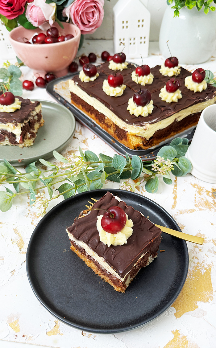 Ein köstliches Blech Donauwelle-Kuchen, dekoriert mit Sahnetupfern und frischen Kirschen. Ein einzelnes Stück des Kuchens liegt auf einem schwarzen Teller mit einer goldenen Gabel. Im Hintergrund befinden sich eine Schüssel voller Kirschen, rosafarbene Blumen und kleine weiße Dekohäuschen. Der Kuchen zeigt die typischen Schichten von hellem und dunklem Teig, Buttercreme und einer Schokoladenglasur