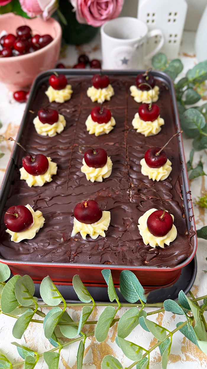 Ein köstliches Blech Donauwelle-Kuchen, dekoriert mit Sahnetupfern und frischen Kirschen. Ein einzelnes Stück des Kuchens liegt auf einem schwarzen Teller mit einer goldenen Gabel. Im Hintergrund befinden sich eine Schüssel voller Kirschen, rosafarbene Blumen und kleine weiße Dekohäuschen. Der Kuchen zeigt die typischen Schichten von hellem und dunklem Teig, Buttercreme und einer Schokoladenglasur