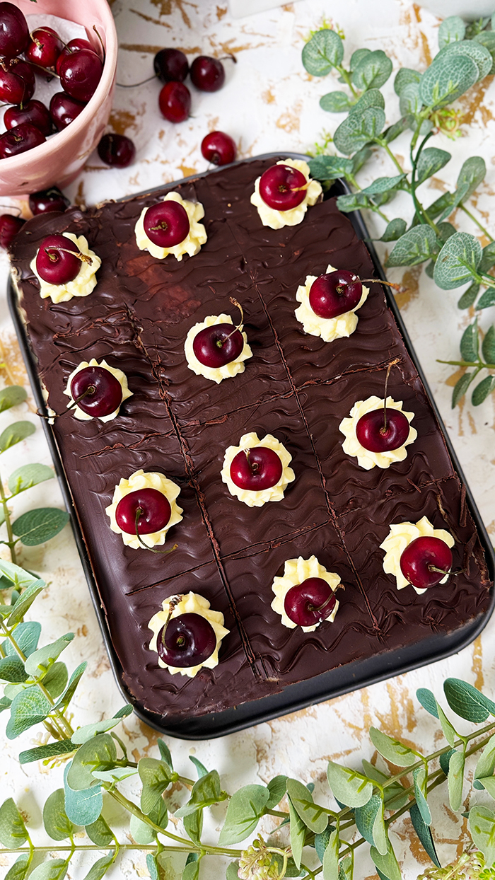 Ein köstliches Blech Donauwelle-Kuchen, dekoriert mit Sahnetupfern und frischen Kirschen. Ein einzelnes Stück des Kuchens liegt auf einem schwarzen Teller mit einer goldenen Gabel. Im Hintergrund befinden sich eine Schüssel voller Kirschen, rosafarbene Blumen und kleine weiße Dekohäuschen. Der Kuchen zeigt die typischen Schichten von hellem und dunklem Teig, Buttercreme und einer Schokoladenglasur