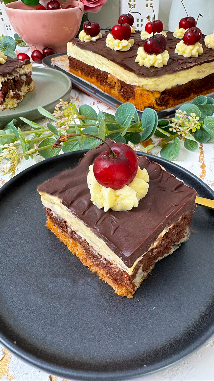 Ein köstliches Blech Donauwelle-Kuchen, dekoriert mit Sahnetupfern und frischen Kirschen. Ein einzelnes Stück des Kuchens liegt auf einem schwarzen Teller mit einer goldenen Gabel. Im Hintergrund befinden sich eine Schüssel voller Kirschen, rosafarbene Blumen und kleine weiße Dekohäuschen. Der Kuchen zeigt die typischen Schichten von hellem und dunklem Teig, Buttercreme und einer Schokoladenglasur