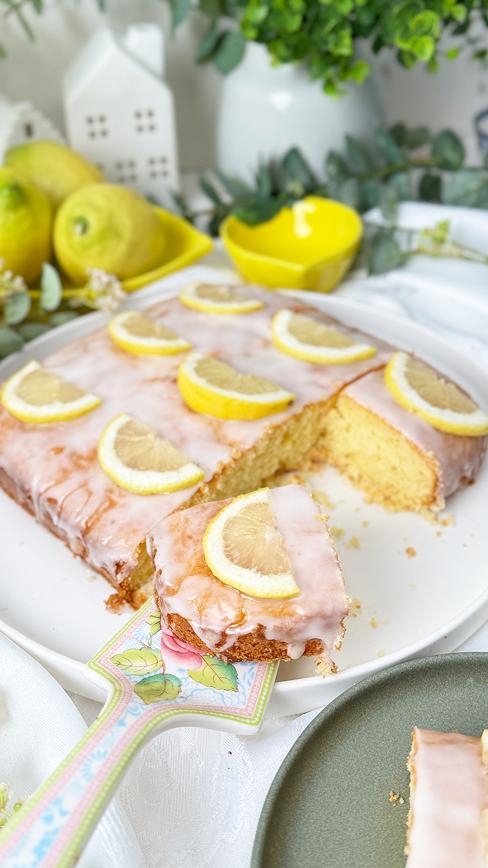 Einfacher Zitronenkuchen vom Blech mit Zuckerguss und Zitronenscheiben dekoriert. Ein Stück auf einem Teller mit einer goldenen Gabel im Vordergrund.