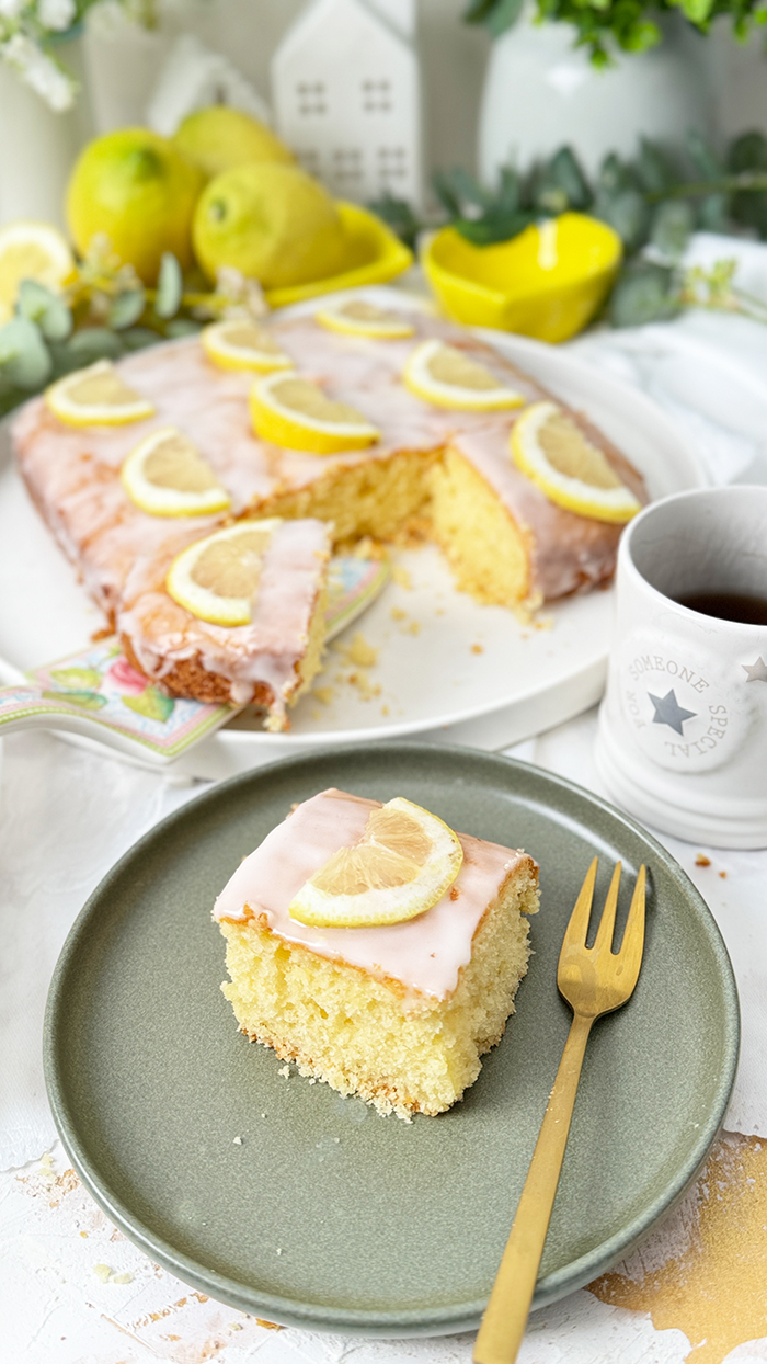 Einfacher Zitronenkuchen vom Blech mit Zuckerguss und Zitronenscheiben dekoriert. Ein Stück auf einem Teller mit einer goldenen Gabel im Vordergrund.