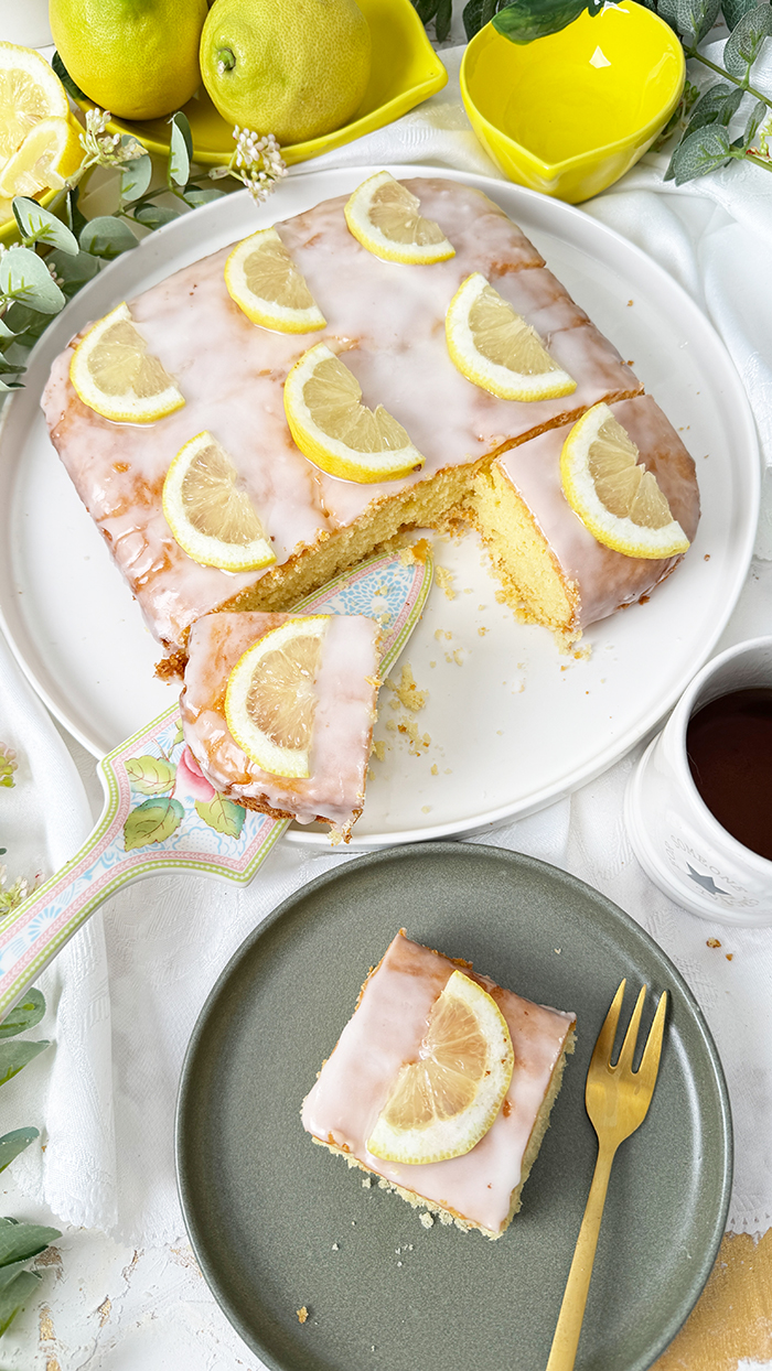 Einfacher Zitronenkuchen vom Blech mit Zuckerguss und Zitronenscheiben dekoriert. Ein Stück auf einem Teller mit einer goldenen Gabel im Vordergrund.