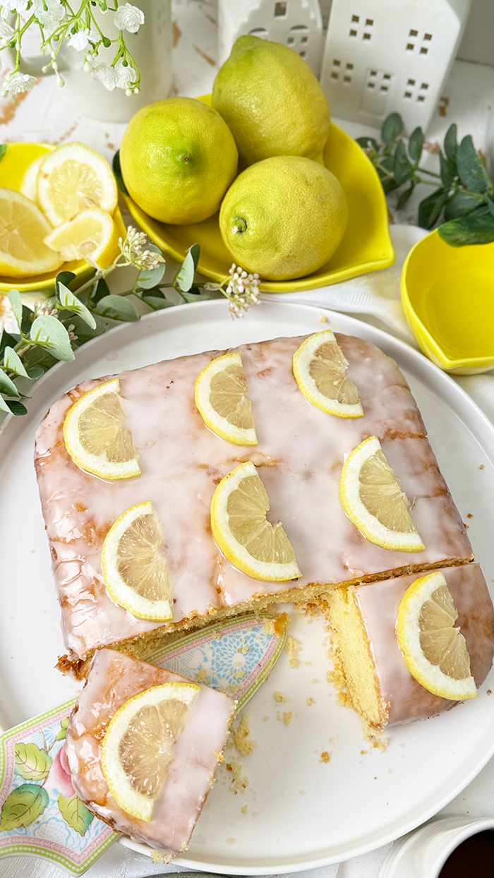 Einfacher Zitronenkuchen vom Blech mit Zuckerguss und Zitronenscheiben dekoriert. Ein Stück auf einem Teller mit einer goldenen Gabel im Vordergrund.