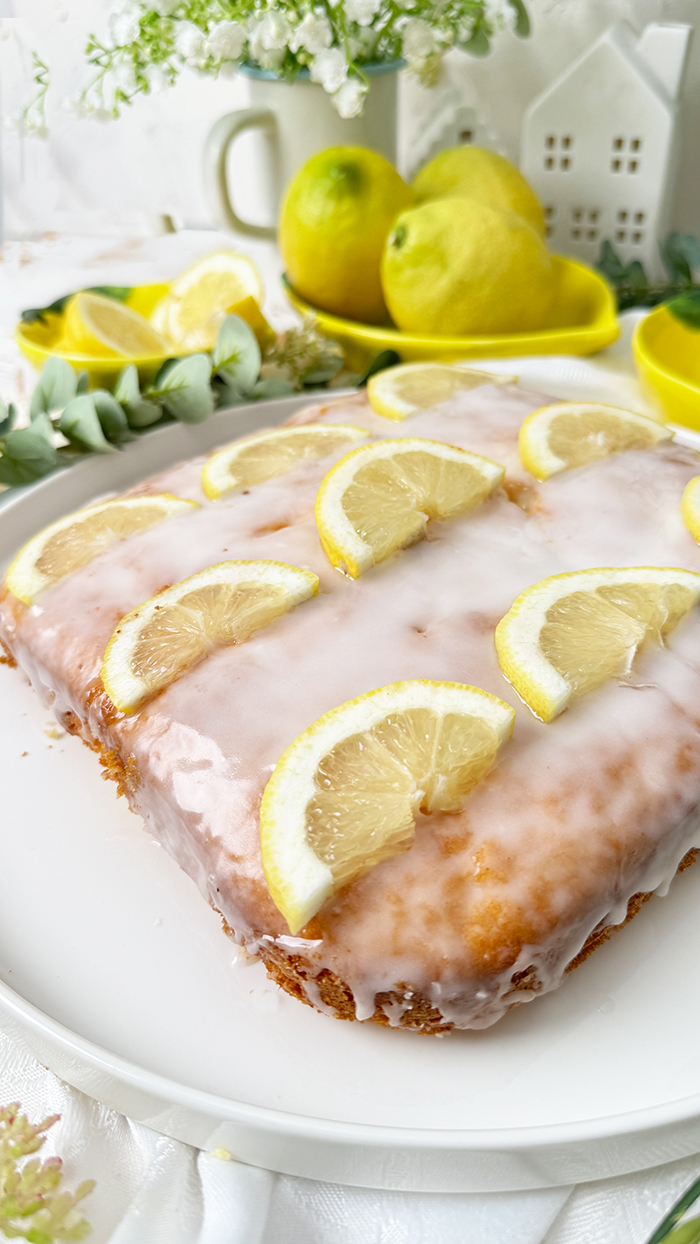 Einfacher Zitronenkuchen vom Blech mit Zuckerguss und Zitronenscheiben dekoriert. Ein Stück auf einem Teller mit einer goldenen Gabel im Vordergrund.