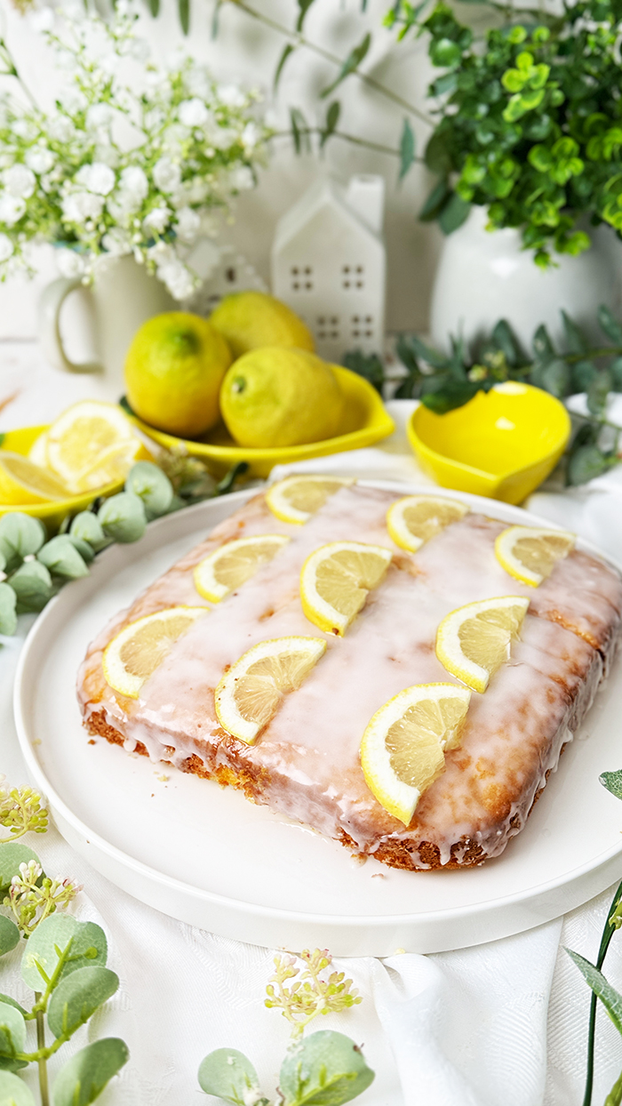 Einfacher Zitronenkuchen vom Blech mit Zuckerguss und Zitronenscheiben dekoriert. Ein Stück auf einem Teller mit einer goldenen Gabel im Vordergrund.