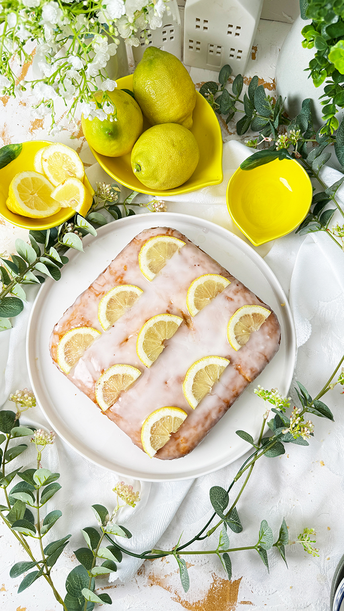 Einfacher Zitronenkuchen vom Blech mit Zuckerguss und Zitronenscheiben dekoriert. Ein Stück auf einem Teller mit einer goldenen Gabel im Vordergrund.