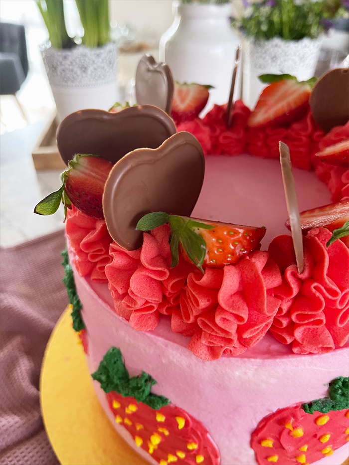 Dies ist ein Bild von einer dekorierten Erdbeer-Schoko-Torte. Die Torte hat eine Basis aus rosa Creme und zeigt mehrere handgefertigte Erdbeerdesigns an den Seiten. Der obere Teil der Torte ist mit rotem Rüschen-Creme, frischen Erdbeeren und Schokoladendekorationen in Herzform verziert. Die Torte steht auf einem goldfarbenen Tortenbrett.