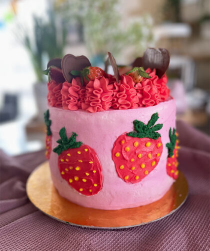 Dies ist ein Bild von einer dekorierten Erdbeer-Schoko-Torte. Die Torte hat eine Basis aus rosa Creme und zeigt mehrere handgefertigte Erdbeerdesigns an den Seiten. Der obere Teil der Torte ist mit rotem Rüschen-Creme, frischen Erdbeeren und Schokoladendekorationen in Herzform verziert. Die Torte steht auf einem goldfarbenen Tortenbrett.