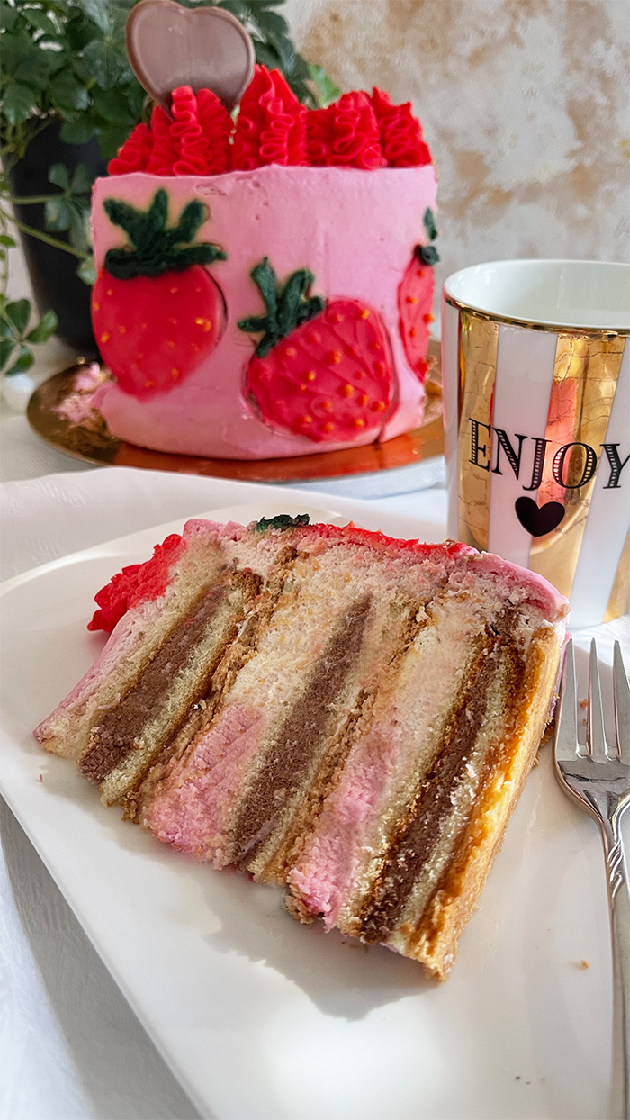 Dies ist ein Bild von einer dekorierten Erdbeer-Schoko-Torte. Die Torte hat eine Basis aus rosa Creme und zeigt mehrere handgefertigte Erdbeerdesigns an den Seiten. Der obere Teil der Torte ist mit rotem Rüschen-Creme, frischen Erdbeeren und Schokoladendekorationen in Herzform verziert. Die Torte steht auf einem goldfarbenen Tortenbrett.