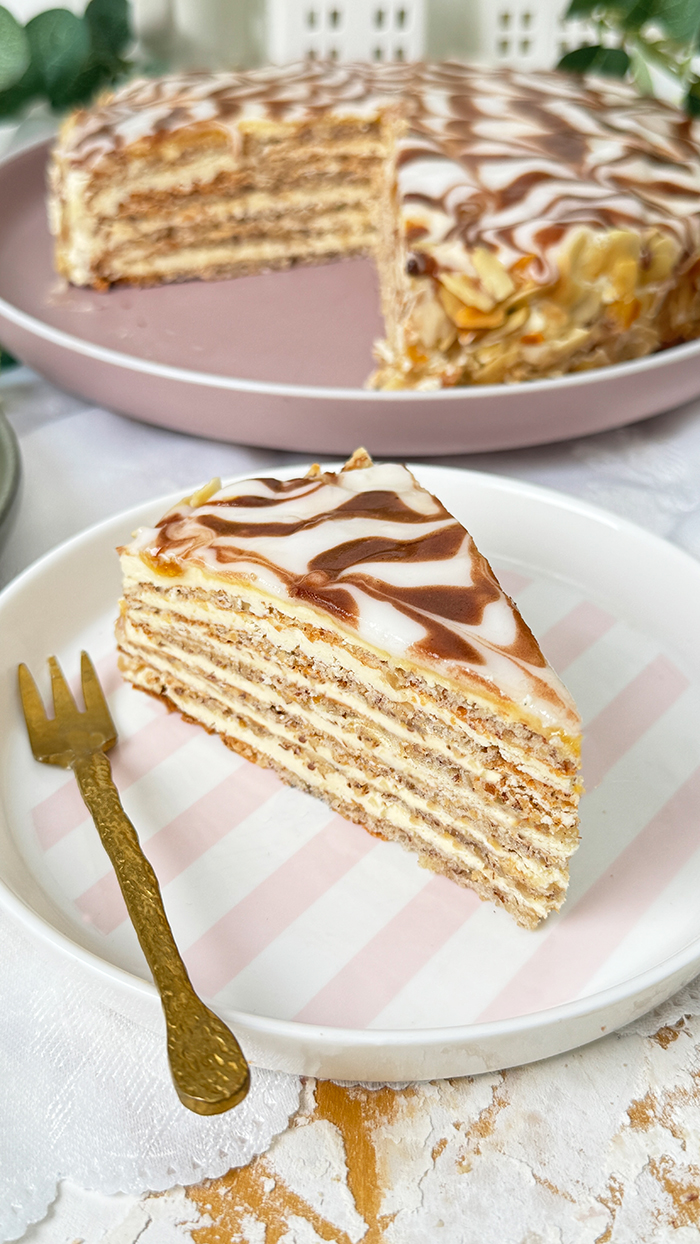 Das Bild zeigt eine Esterházy-Torte, die aus mehreren dünnen Schichten besteht, gefüllt mit Buttercreme, und oben mit einer weißen Glasur und dem typischen Kakaomuster verziert ist.