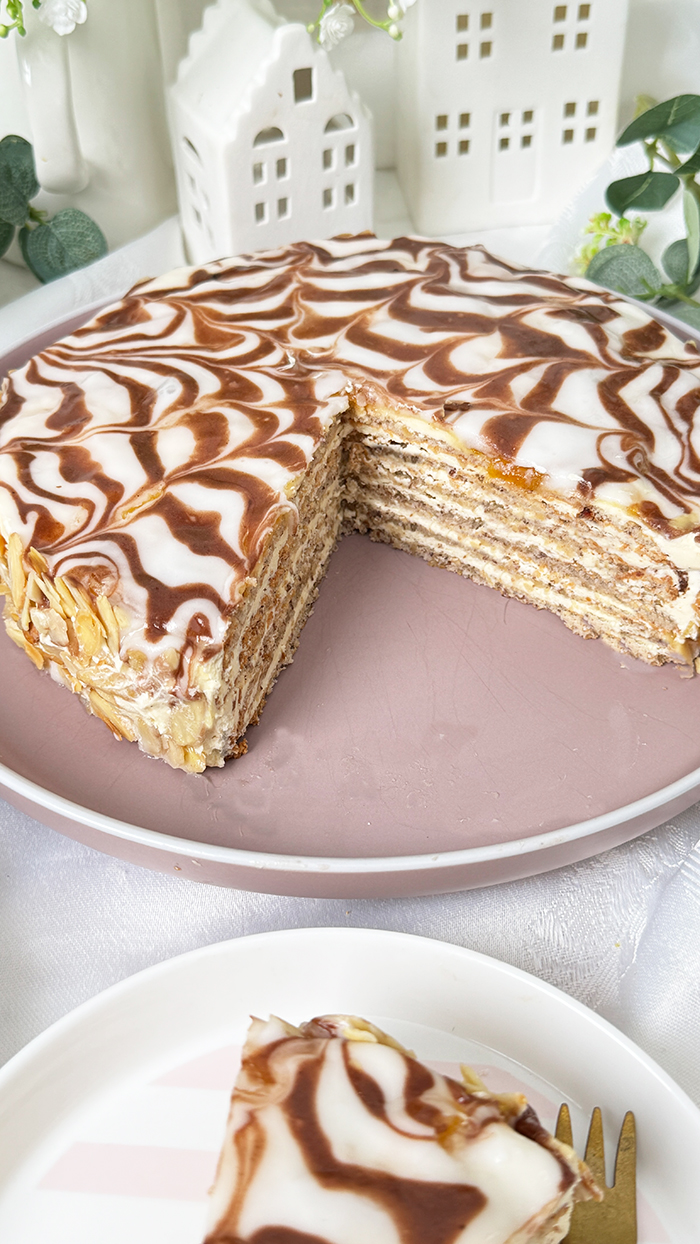 Das Bild zeigt eine Esterházy-Torte, die aus mehreren dünnen Schichten besteht, gefüllt mit Buttercreme, und oben mit einer weißen Glasur und dem typischen Kakaomuster verziert ist.