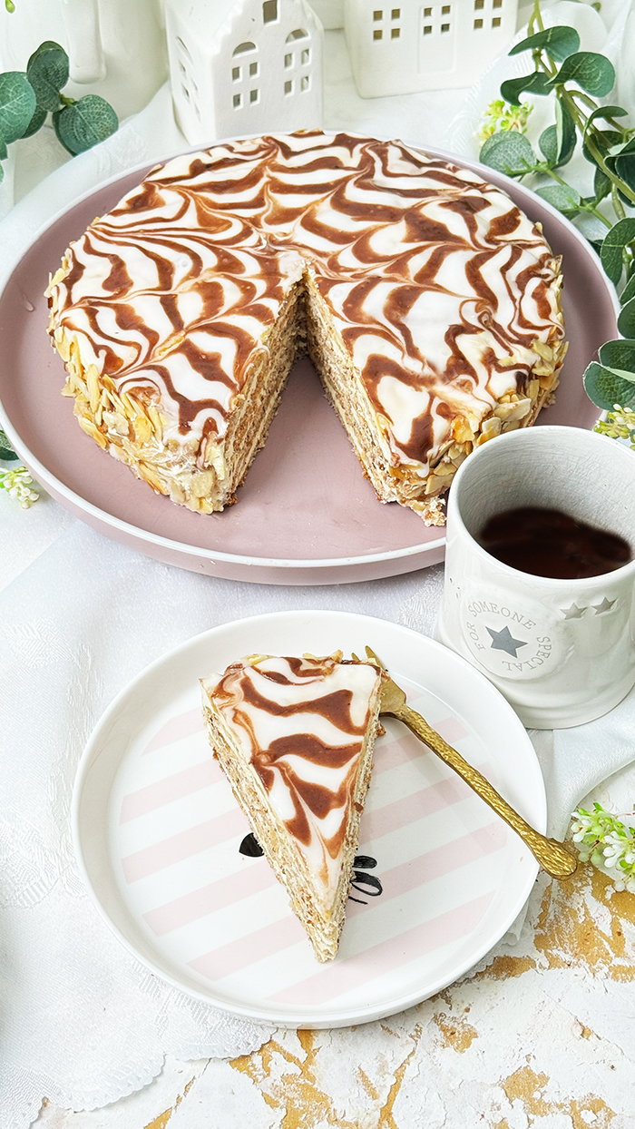 Das Bild zeigt eine Esterházy-Torte, die aus mehreren dünnen Schichten besteht, gefüllt mit Buttercreme, und oben mit einer weißen Glasur und dem typischen Kakaomuster verziert ist.