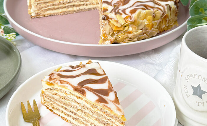 Das Bild zeigt eine Esterházy-Torte, die aus mehreren dünnen Schichten besteht, gefüllt mit Buttercreme, und oben mit einer weißen Glasur und dem typischen Kakaomuster verziert ist.