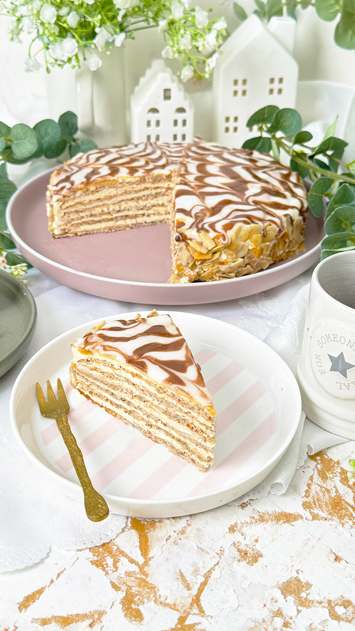 Das Bild zeigt eine Esterházy-Torte, die aus mehreren dünnen Schichten besteht, gefüllt mit Buttercreme, und oben mit einer weißen Glasur und dem typischen Kakaomuster verziert ist.