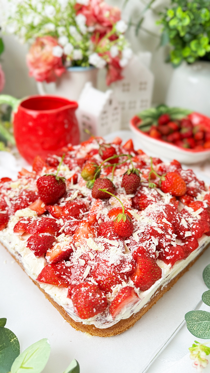 Das Bild zeigt einen Fantakuchen, der auf einer weißen Platte serviert wird. Der Kuchen hat eine rechteckige Form und ist mit einer Schicht Sahnecreme überzogen. Darauf befinden sich zahlreiche frische Erdbeeren, die in Stücke geschnitten sind und einige ganze Erdbeeren mit Stielen als Dekoration. Außerdem sind weiße Schokoladenraspeln über die Erdbeeren gestreut. Im Hintergrund sind unscharf Pflanzen, Blumen und weitere Erdbeeren in einer Schale zu sehen, die eine frische und einladende Atmosphäre schaffen.