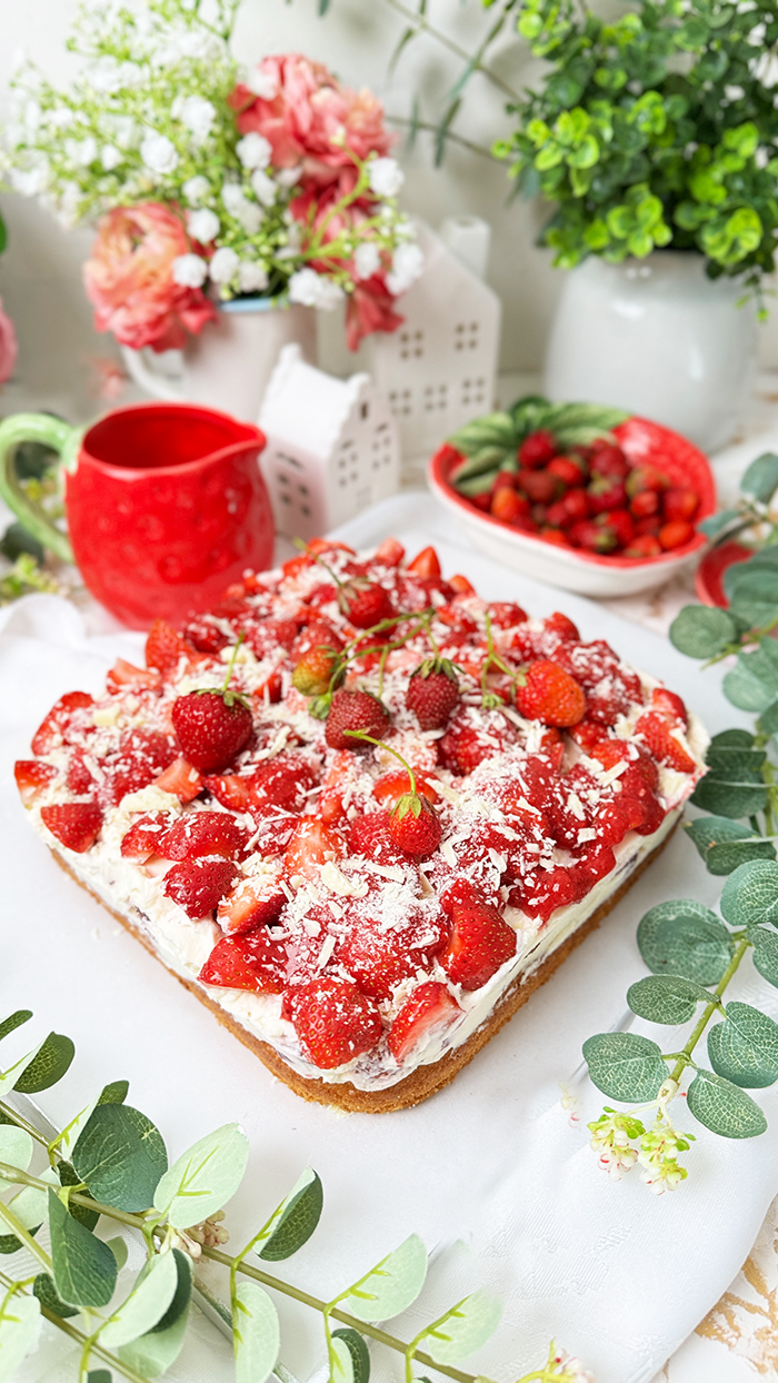 Das Bild zeigt einen Fantakuchen, der auf einer weißen Platte serviert wird. Der Kuchen hat eine rechteckige Form und ist mit einer Schicht Sahnecreme überzogen. Darauf befinden sich zahlreiche frische Erdbeeren, die in Stücke geschnitten sind und einige ganze Erdbeeren mit Stielen als Dekoration. Außerdem sind weiße Schokoladenraspeln über die Erdbeeren gestreut. Im Hintergrund sind unscharf Pflanzen, Blumen und weitere Erdbeeren in einer Schale zu sehen, die eine frische und einladende Atmosphäre schaffen.