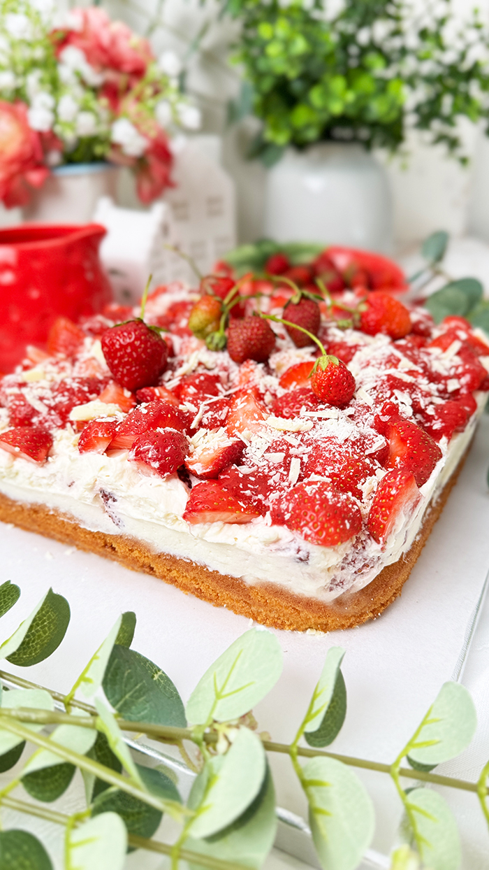 Das Bild zeigt einen Fantakuchen, der auf einer weißen Platte serviert wird. Der Kuchen hat eine rechteckige Form und ist mit einer Schicht Sahnecreme überzogen. Darauf befinden sich zahlreiche frische Erdbeeren, die in Stücke geschnitten sind und einige ganze Erdbeeren mit Stielen als Dekoration. Außerdem sind weiße Schokoladenraspeln über die Erdbeeren gestreut. Im Hintergrund sind unscharf Pflanzen, Blumen und weitere Erdbeeren in einer Schale zu sehen, die eine frische und einladende Atmosphäre schaffen.