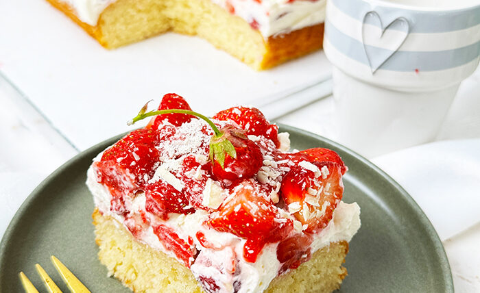 Das Bild zeigt einen Fantakuchen, der auf einer weißen Platte serviert wird. Der Kuchen hat eine rechteckige Form und ist mit einer Schicht Sahnecreme überzogen. Darauf befinden sich zahlreiche frische Erdbeeren, die in Stücke geschnitten sind und einige ganze Erdbeeren mit Stielen als Dekoration. Außerdem sind weiße Schokoladenraspeln über die Erdbeeren gestreut. Im Hintergrund sind unscharf Pflanzen, Blumen und weitere Erdbeeren in einer Schale zu sehen, die eine frische und einladende Atmosphäre schaffen.