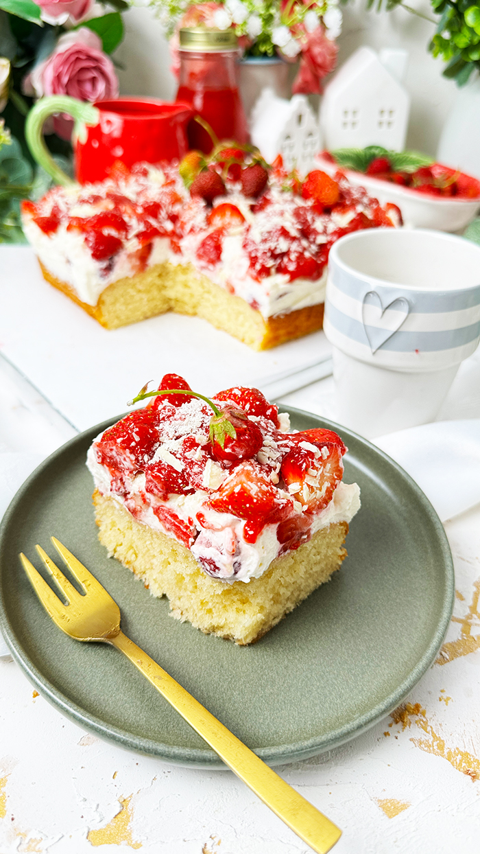 Das Bild zeigt einen Fantakuchen, der auf einer weißen Platte serviert wird. Der Kuchen hat eine rechteckige Form und ist mit einer Schicht Sahnecreme überzogen. Darauf befinden sich zahlreiche frische Erdbeeren, die in Stücke geschnitten sind und einige ganze Erdbeeren mit Stielen als Dekoration. Außerdem sind weiße Schokoladenraspeln über die Erdbeeren gestreut. Im Hintergrund sind unscharf Pflanzen, Blumen und weitere Erdbeeren in einer Schale zu sehen, die eine frische und einladende Atmosphäre schaffen.