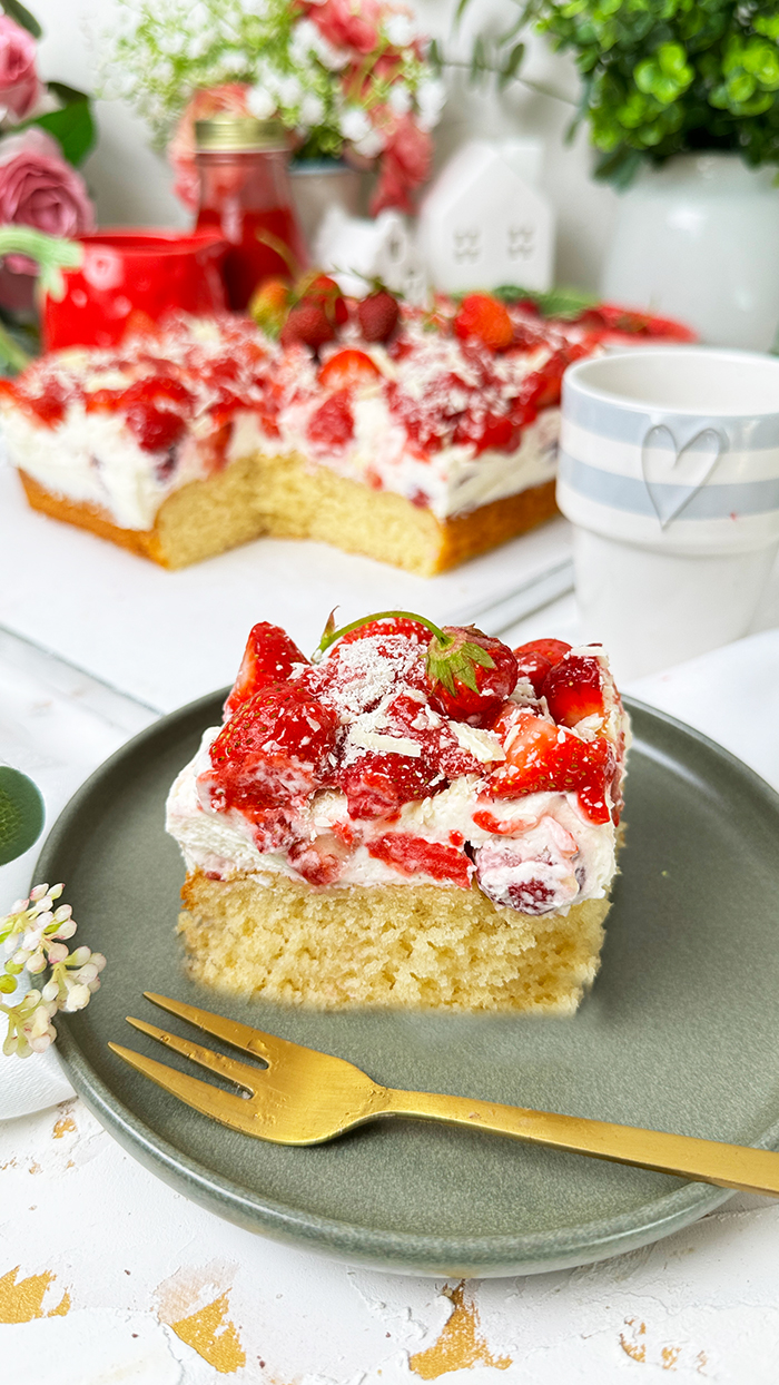 Das Bild zeigt einen Fantakuchen, der auf einer weißen Platte serviert wird. Der Kuchen hat eine rechteckige Form und ist mit einer Schicht Sahnecreme überzogen. Darauf befinden sich zahlreiche frische Erdbeeren, die in Stücke geschnitten sind und einige ganze Erdbeeren mit Stielen als Dekoration. Außerdem sind weiße Schokoladenraspeln über die Erdbeeren gestreut. Im Hintergrund sind unscharf Pflanzen, Blumen und weitere Erdbeeren in einer Schale zu sehen, die eine frische und einladende Atmosphäre schaffen.