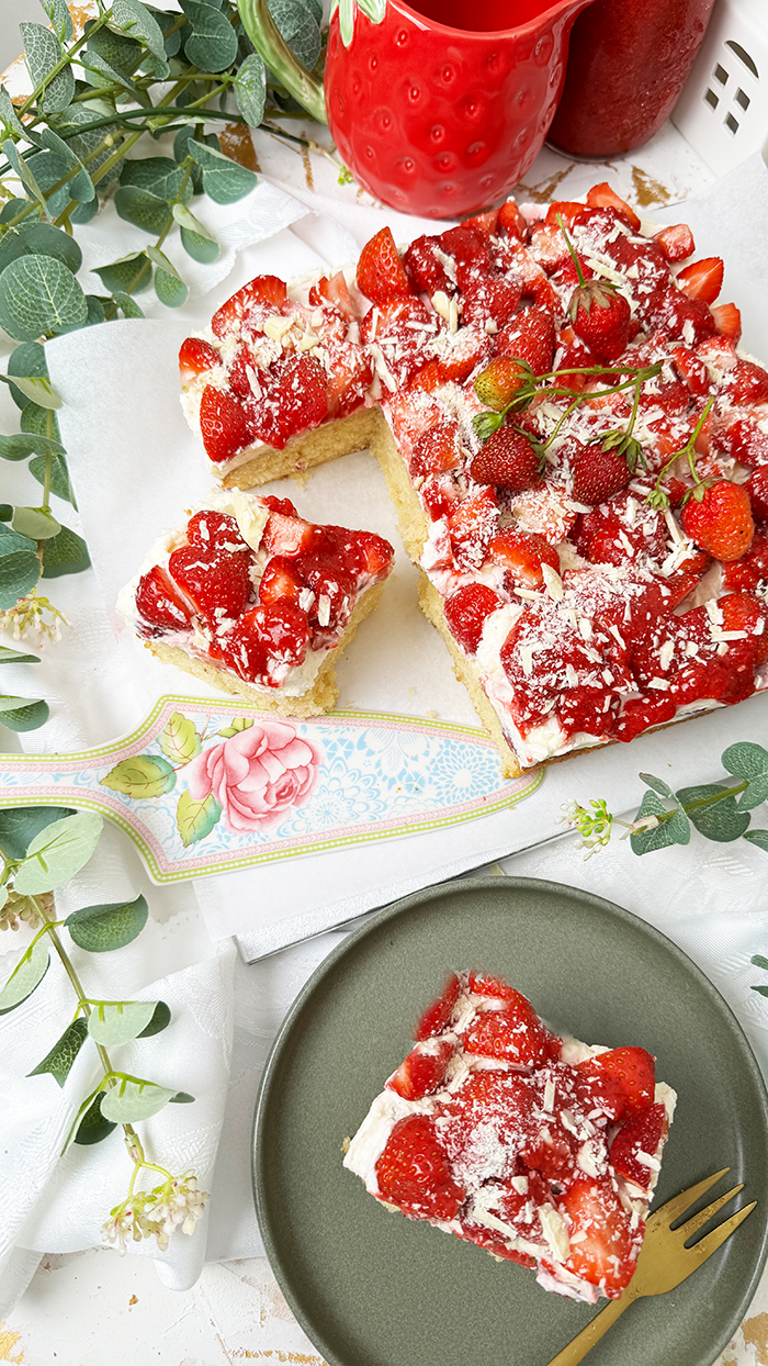 Das Bild zeigt einen Fantakuchen, der auf einer weißen Platte serviert wird. Der Kuchen hat eine rechteckige Form und ist mit einer Schicht Sahnecreme überzogen. Darauf befinden sich zahlreiche frische Erdbeeren, die in Stücke geschnitten sind und einige ganze Erdbeeren mit Stielen als Dekoration. Außerdem sind weiße Schokoladenraspeln über die Erdbeeren gestreut. Im Hintergrund sind unscharf Pflanzen, Blumen und weitere Erdbeeren in einer Schale zu sehen, die eine frische und einladende Atmosphäre schaffen.