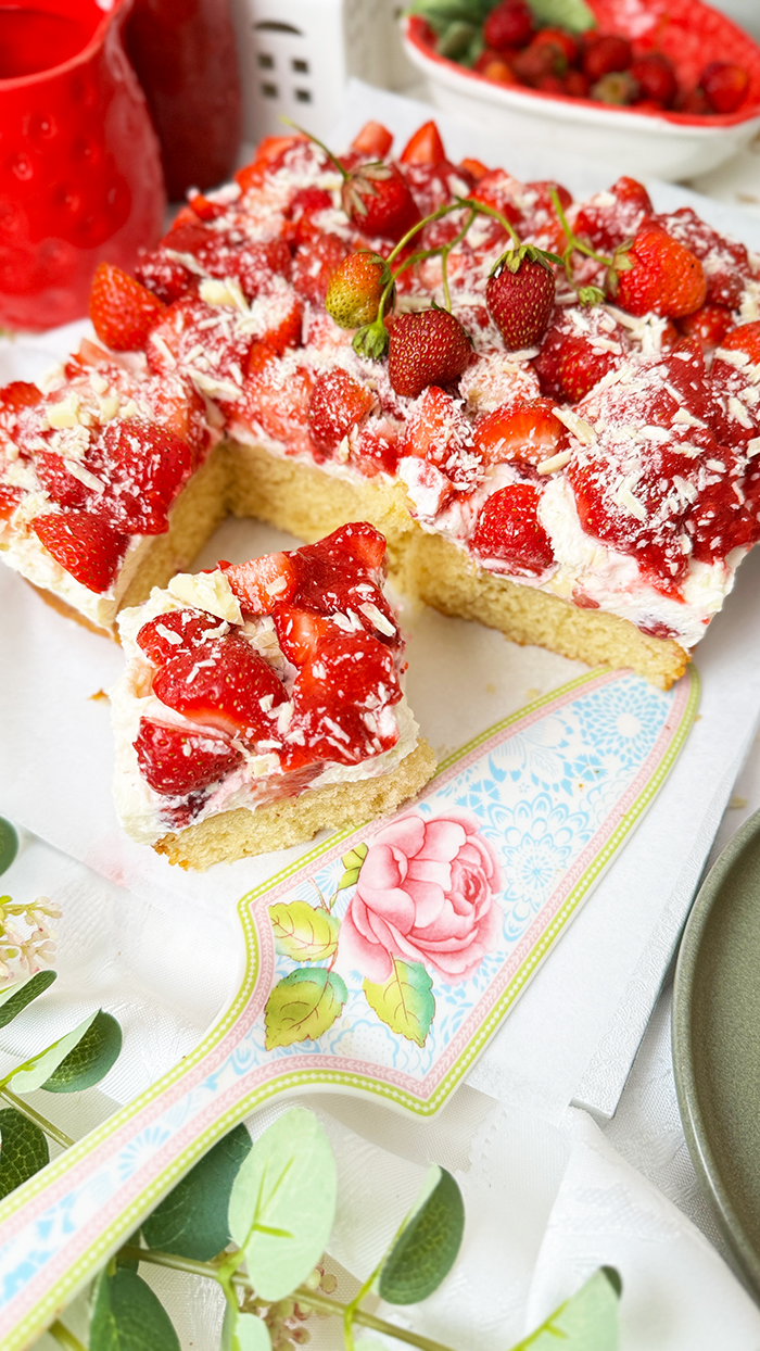 Das Bild zeigt einen Fantakuchen, der auf einer weißen Platte serviert wird. Der Kuchen hat eine rechteckige Form und ist mit einer Schicht Sahnecreme überzogen. Darauf befinden sich zahlreiche frische Erdbeeren, die in Stücke geschnitten sind und einige ganze Erdbeeren mit Stielen als Dekoration. Außerdem sind weiße Schokoladenraspeln über die Erdbeeren gestreut. Im Hintergrund sind unscharf Pflanzen, Blumen und weitere Erdbeeren in einer Schale zu sehen, die eine frische und einladende Atmosphäre schaffen.