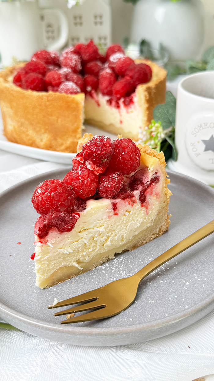 ein Stück Himbeer-Schmand-Kuchen auf einem Teller, garniert mit frischen Himbeeren und leicht mit Puderzucker bestäubt.