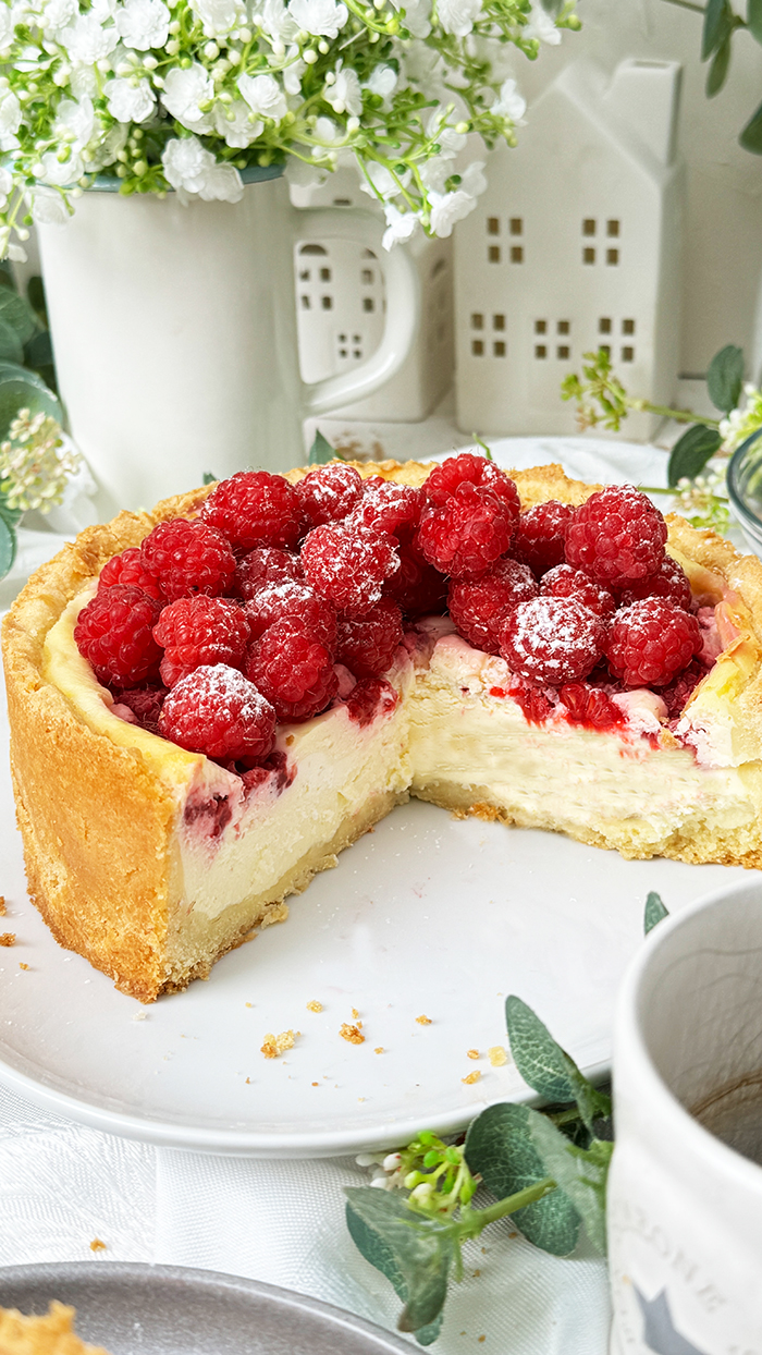 ein Stück Himbeer-Schmand-Kuchen auf einem Teller, garniert mit frischen Himbeeren und leicht mit Puderzucker bestäubt.