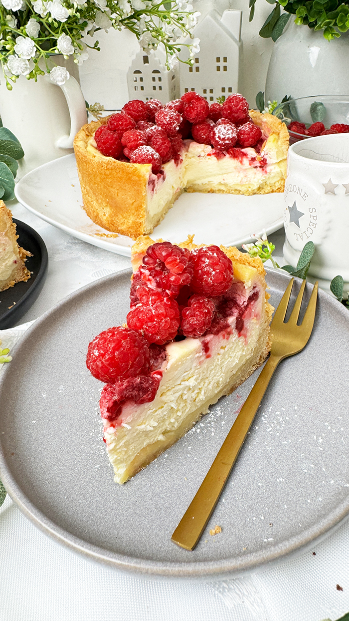 ein Stück Himbeer-Schmand-Kuchen auf einem Teller, garniert mit frischen Himbeeren und leicht mit Puderzucker bestäubt.