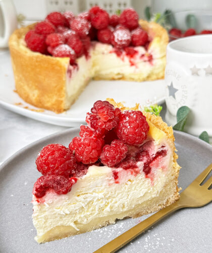 ein Stück Himbeer-Schmand-Kuchen auf einem Teller, garniert mit frischen Himbeeren und leicht mit Puderzucker bestäubt.