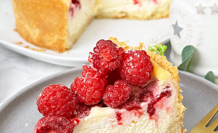 ein Stück Himbeer-Schmand-Kuchen auf einem Teller, garniert mit frischen Himbeeren und leicht mit Puderzucker bestäubt.